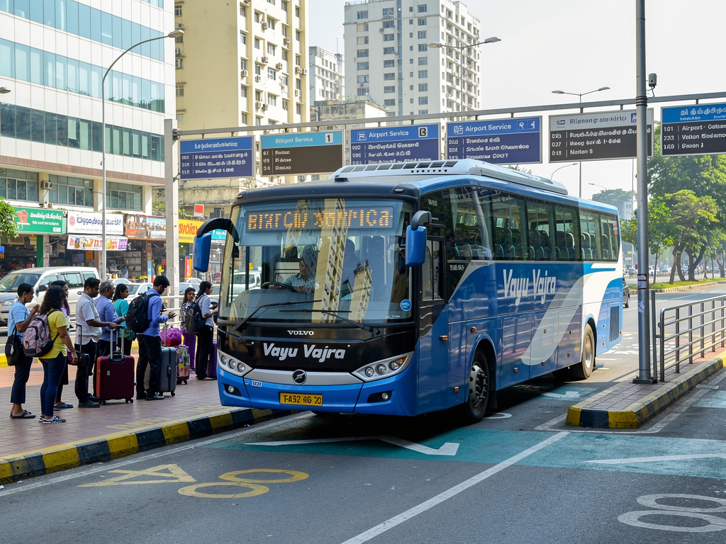 A modern BMTC Vayu Vajra Volvo airport coach parked at a busy urban bus station during daytime, with passengers waiting nearby and city buildings visible in the background, bright natural lighting creating a welcoming atmosphere