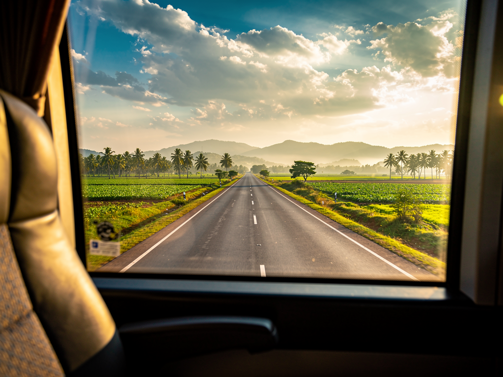 Scenic view from a bus window showing the Bangalore-Mysore highway passing through green agricultural fields with distant hills under a partly cloudy sky